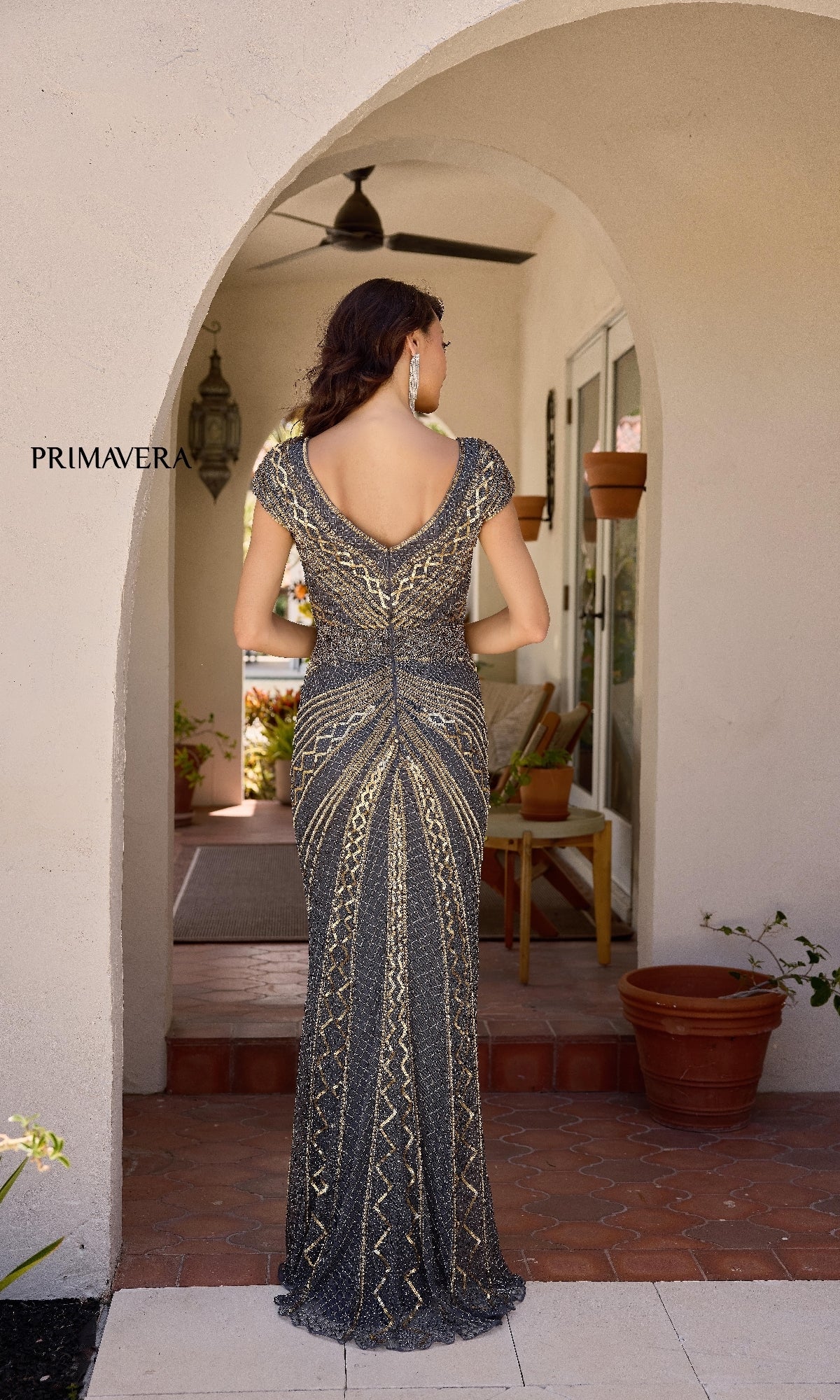A woman in the Primavera 13135 long formal dress, featuring a fitted silhouette and beaded gold and black patterns, stands on a tiled porch under an arched entryway, facing away.